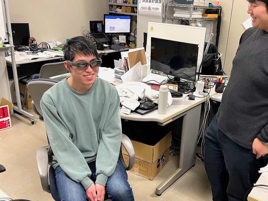 大学生二人が笑いながら映っている。緑色のトレーナーを着用し男子学生が今回のメガネを着用して、座っている。その様子を黒のトレーナーを着用した男子学生が横に立って見ている。